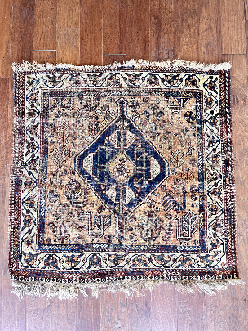 a square antique qashqai rug with a blue medallion, coral field and green accents